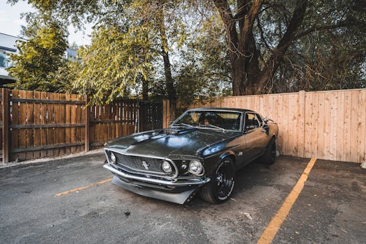 A vintage classic car parked in an outdoor yard surrounded by trees and wooden fences.