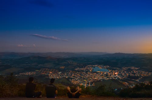 Three Men Sitting on Top of Mountain during Pale Evening Sky While Facing City Full of Lights