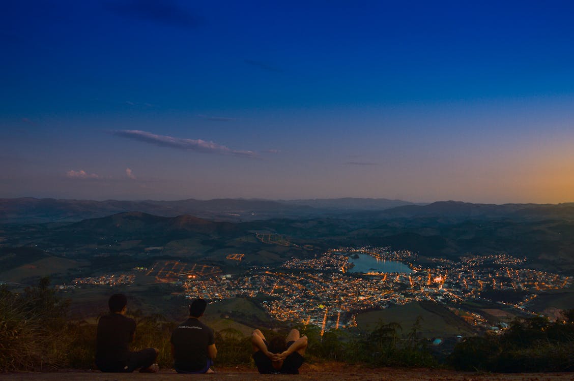 Free Three Men Sitting on Top of Mountain during Pale Evening Sky While Facing City Full of Lights Stock Photo