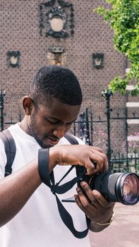A black man in a white shirt adjusts a camera in an urban outdoor setting.