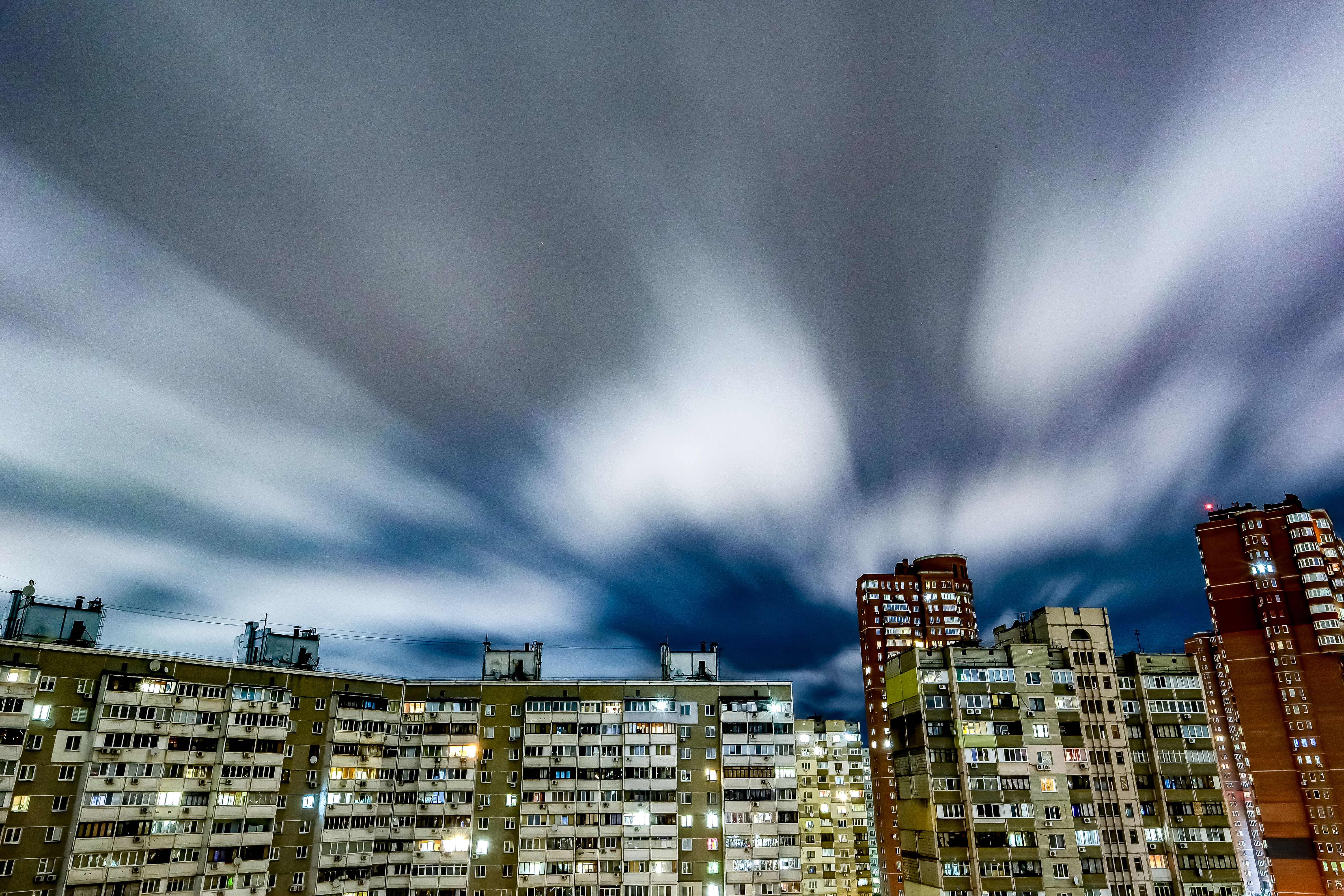 Long exposure of urban skyline with illuminated buildings and fast-moving clouds creating a dynamic city night scene.