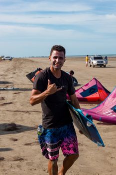 A surfer holding a board and smiling while standing on a beach with kitesurfs in the background.