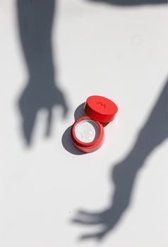 Red skincare ointment container with shadows on white background in sunlight.