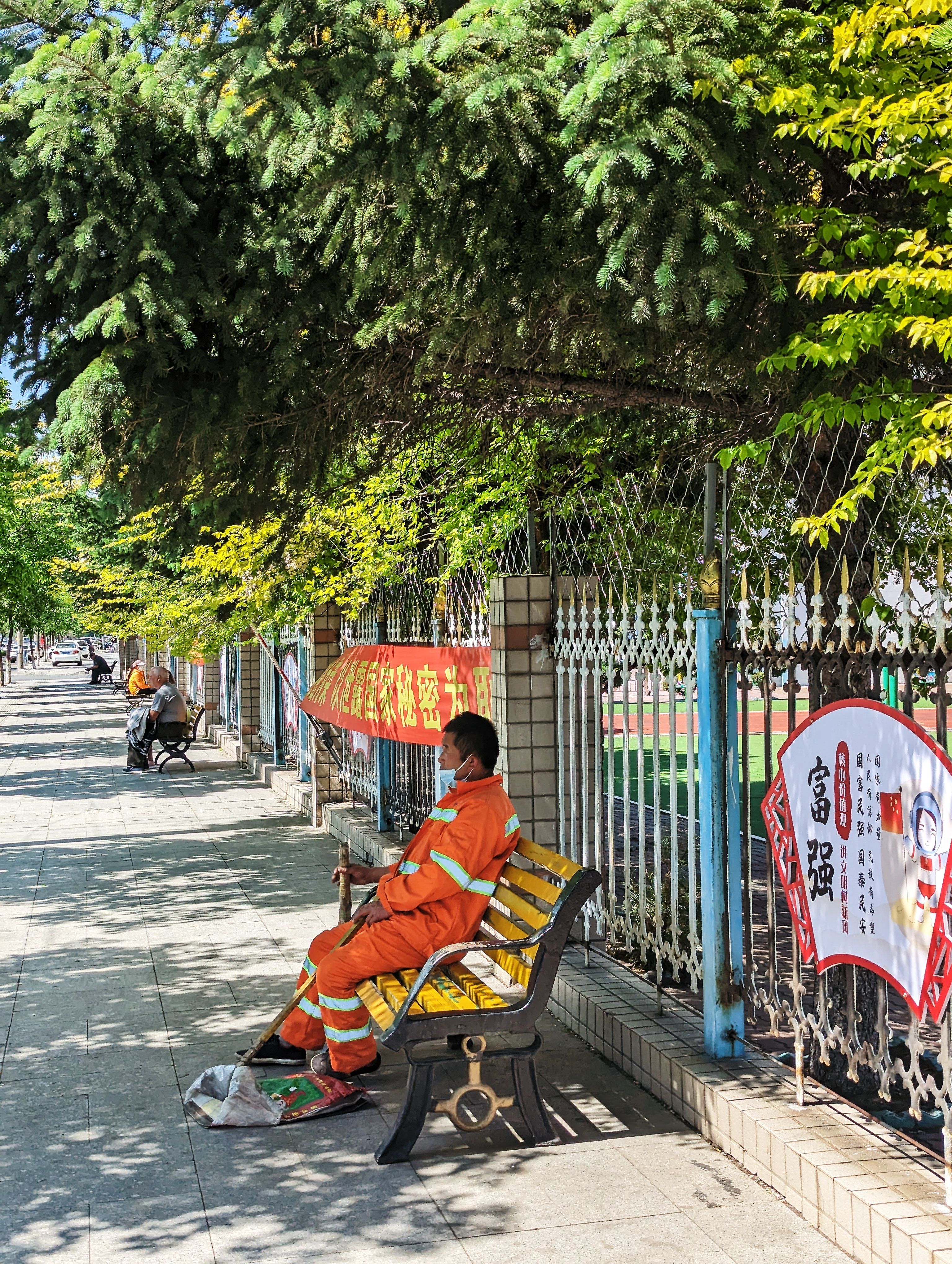 A worker in coveralls takes a break on a bench under trees on a sunny street in Yan Bian, China.