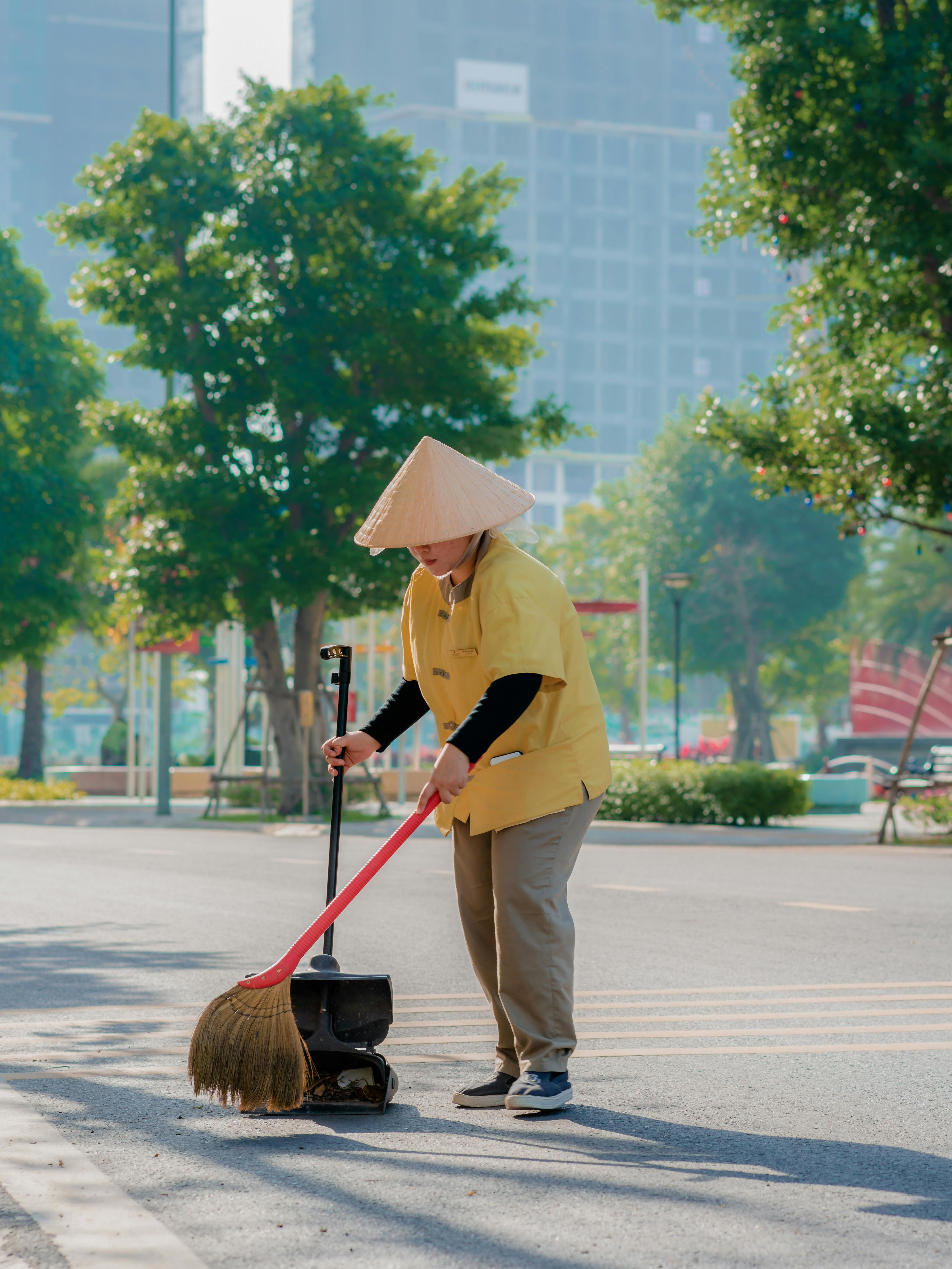 A woman in a conical hat sweeping a city street amidst tall buildings and greenery.