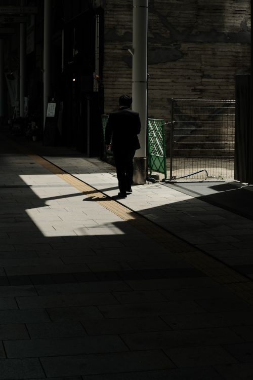 Free A lone man walks down a shadowy urban street, casting a silhouette on the pavement. Stock Photo