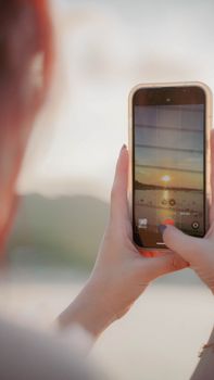 Hands holding a smartphone to capture a stunning sunset on the beach.