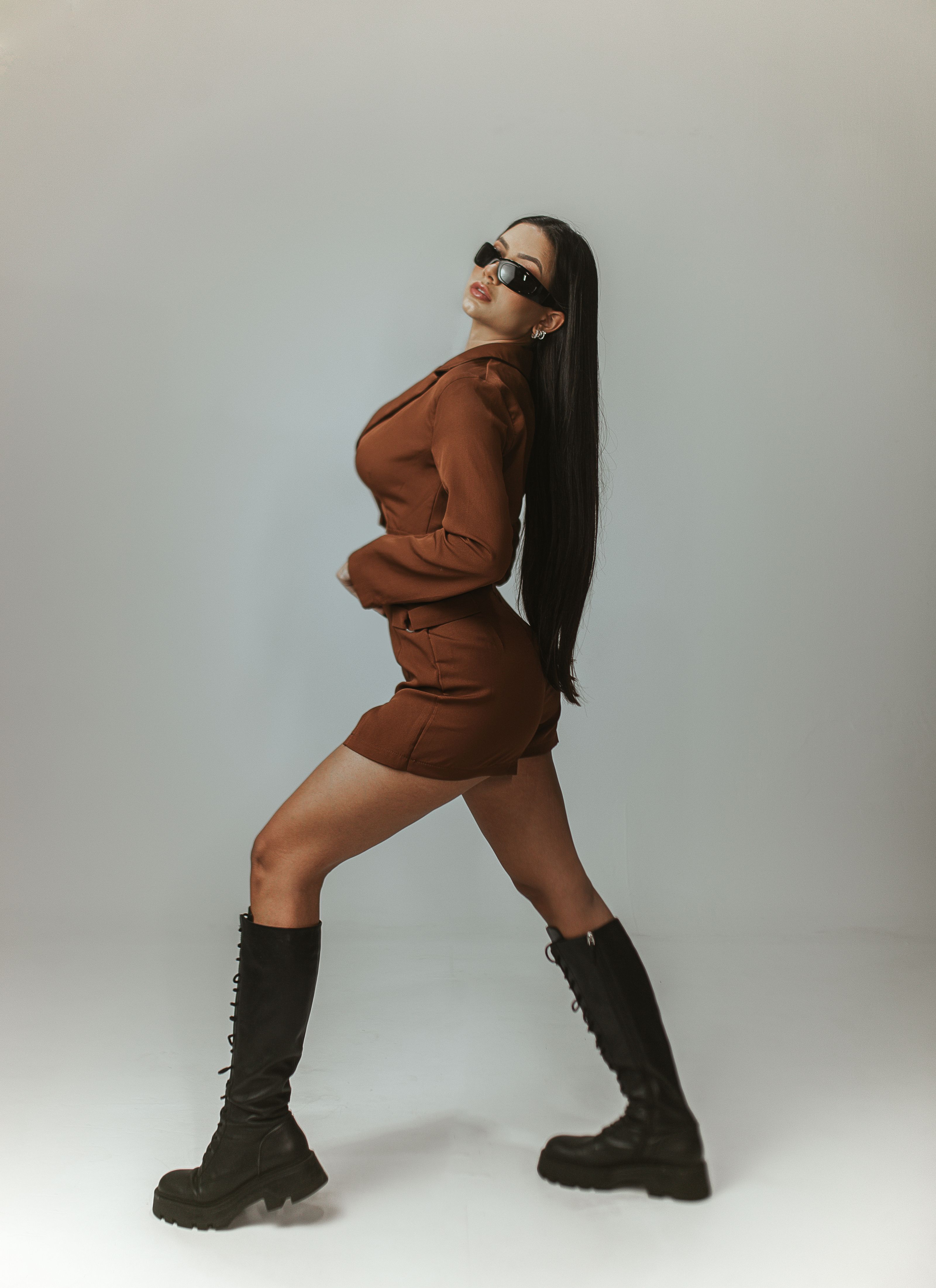 Mulher fashion com cabelo longo posando em um conjunto marrom e botas pretas sobre um fundo cinza.
