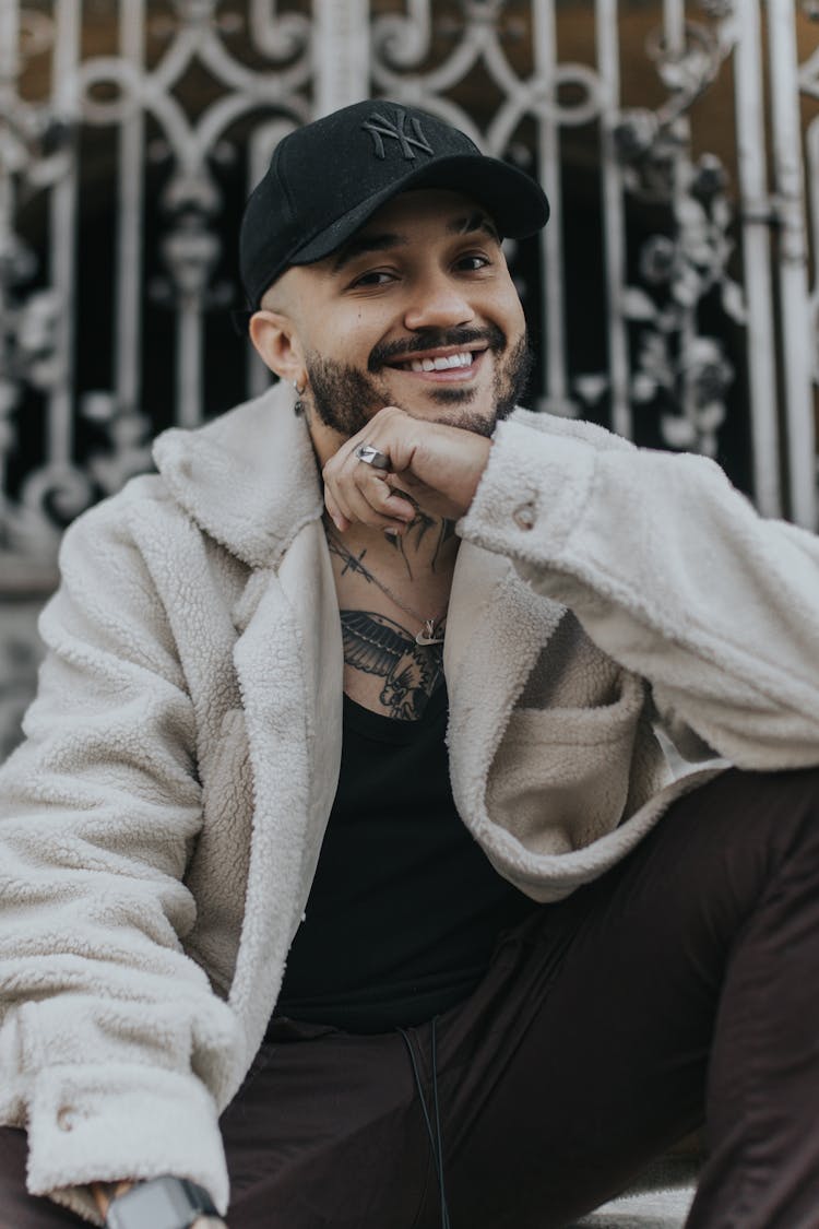 Smiling Man Wearing A Cap Posing By A Metal Gate