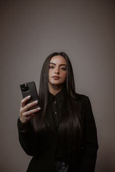 Stylish woman in black clothing confidently holding a smartphone. Studio portrait with grey background.