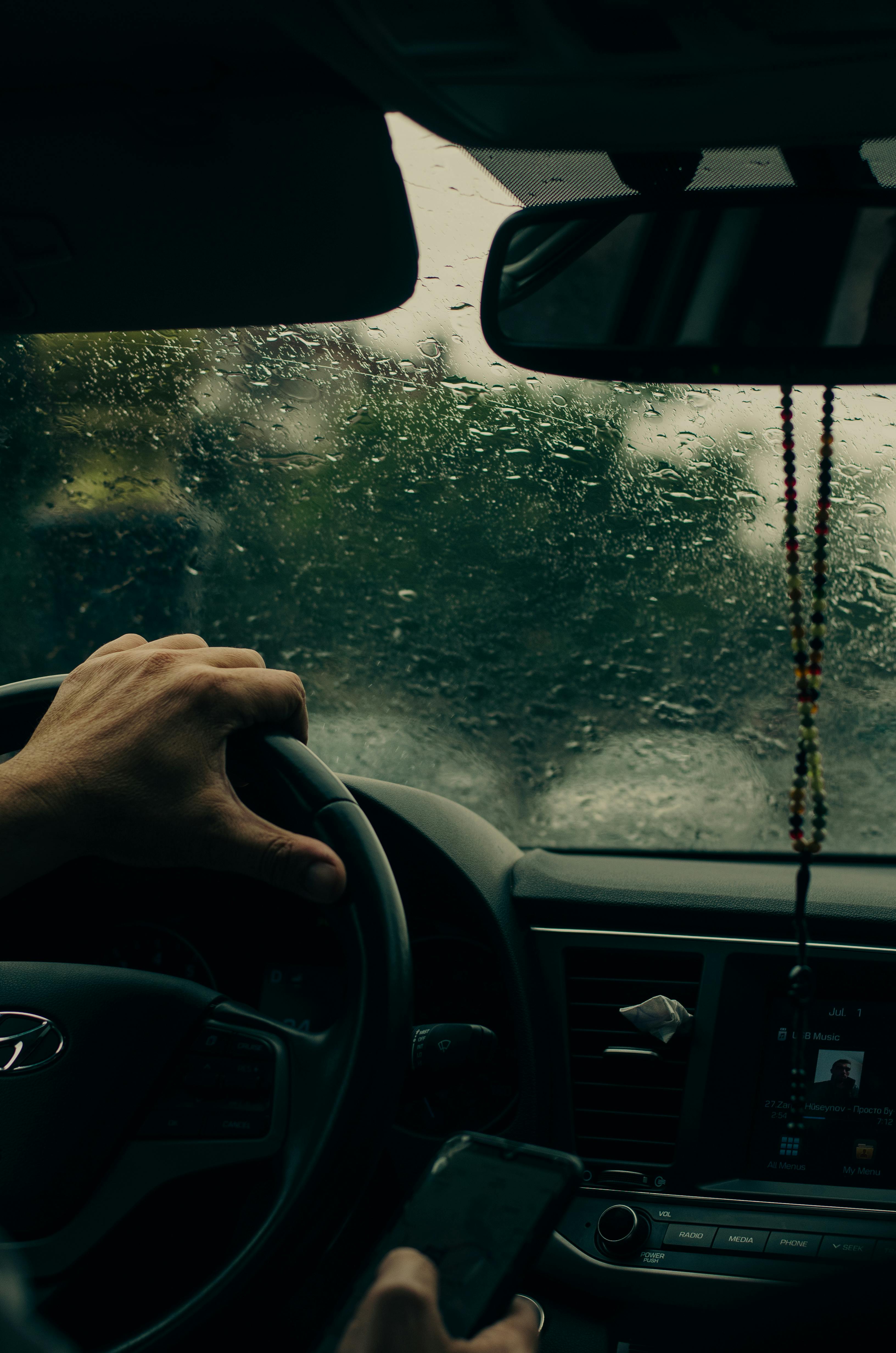 Driver holds a smartphone while steering in a car on a rainy day, emphasizing modern navigation.