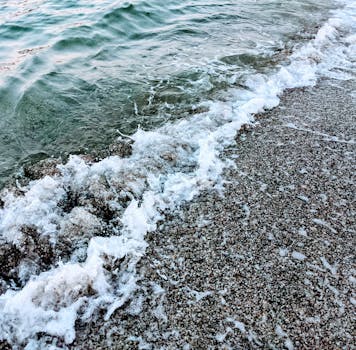 Gentle waves wash over a rocky seashore, creating white foam under a clear sky.