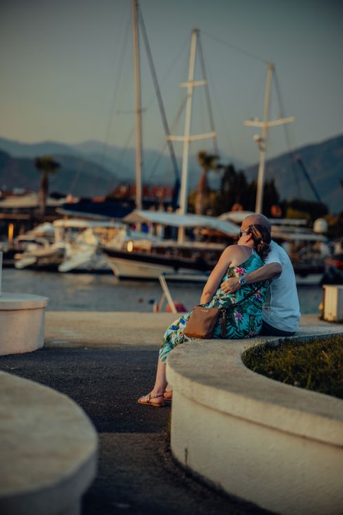 Free A couple sharing an affectionate moment at a marina during a warm summer evening, surrounded by boats and mountains. Stock Photo