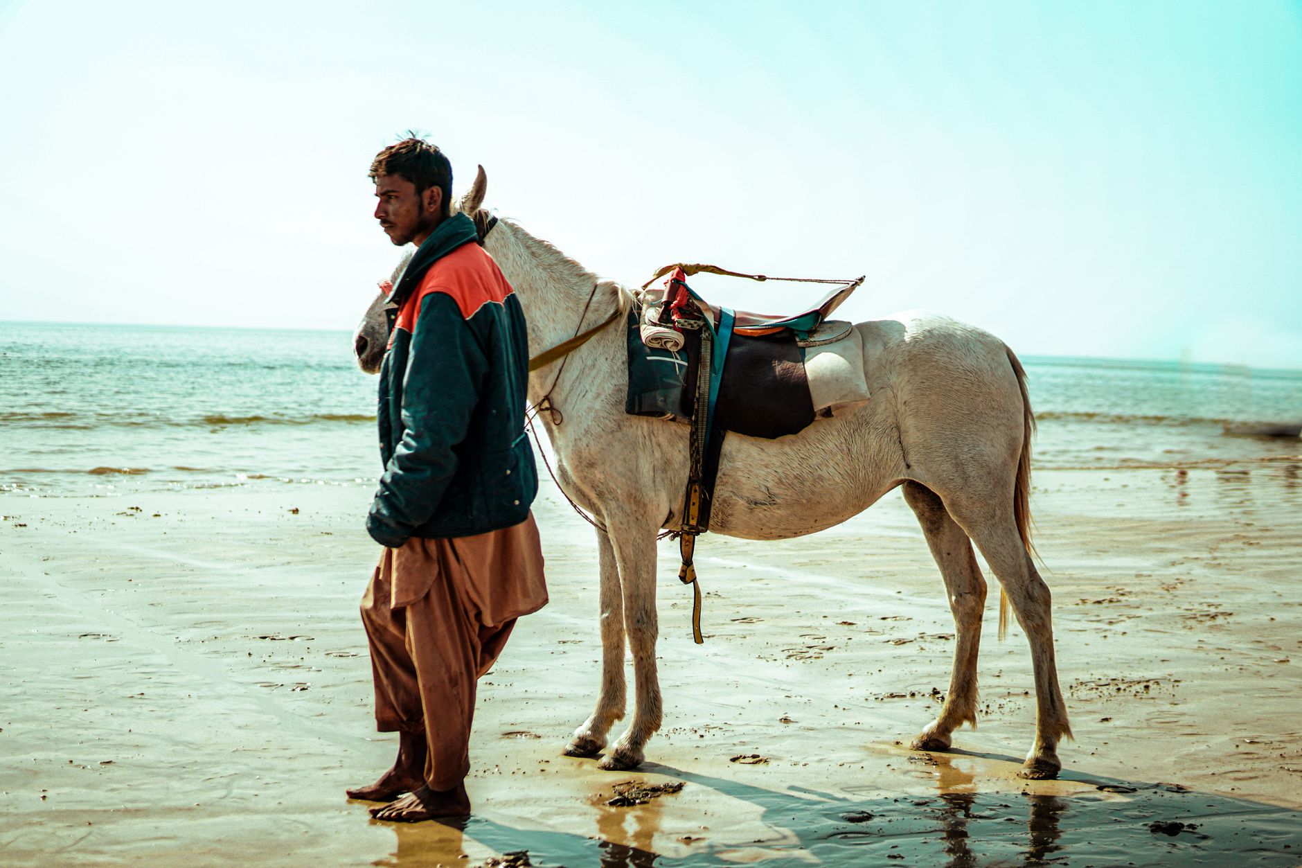 A man stands with a saddled horse on a scenic Karachi beach.