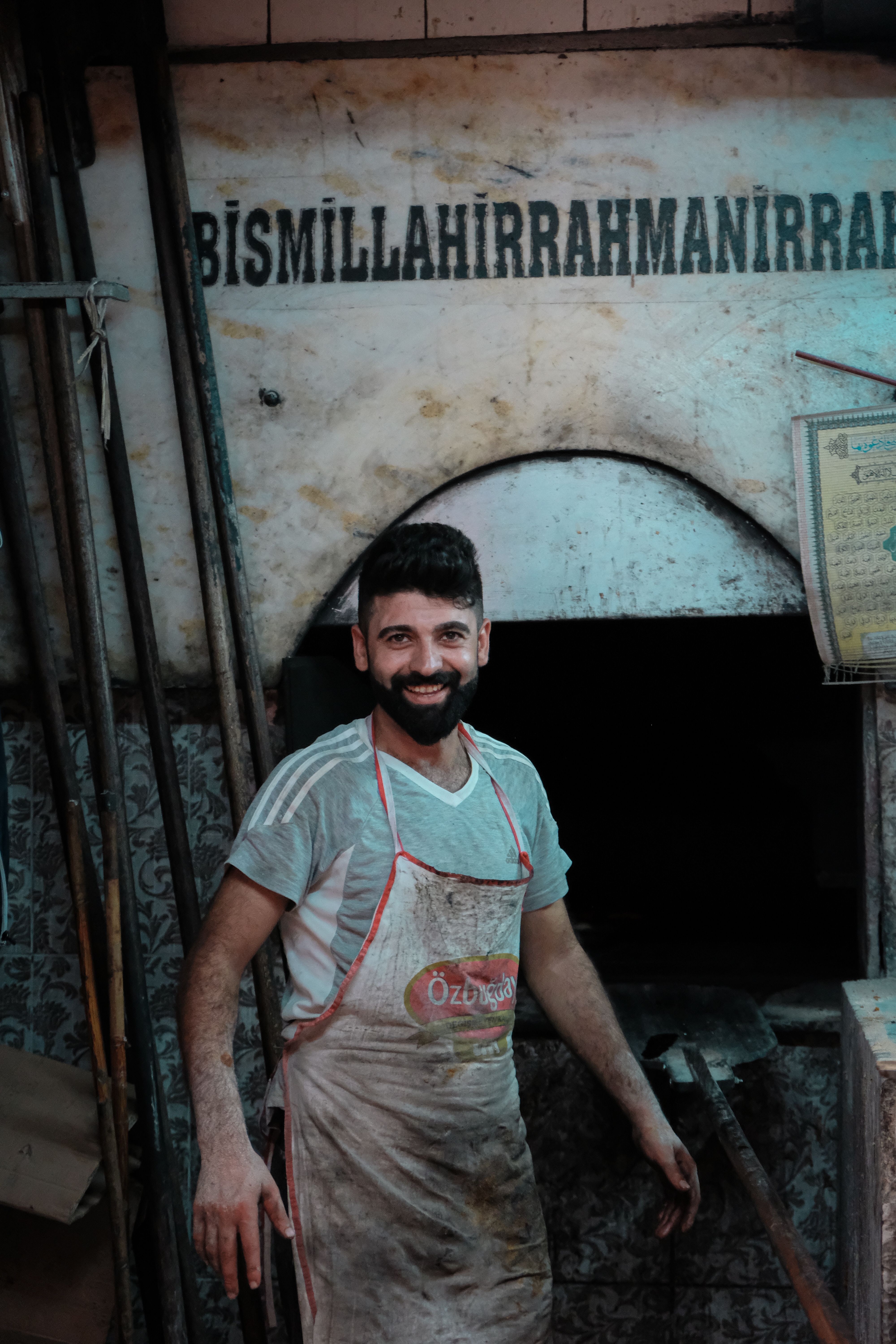 A cheerful baker poses by a traditional oven in an indoor bakery setting.