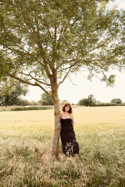 A woman in a black dress stands against a tree in a sunny field.