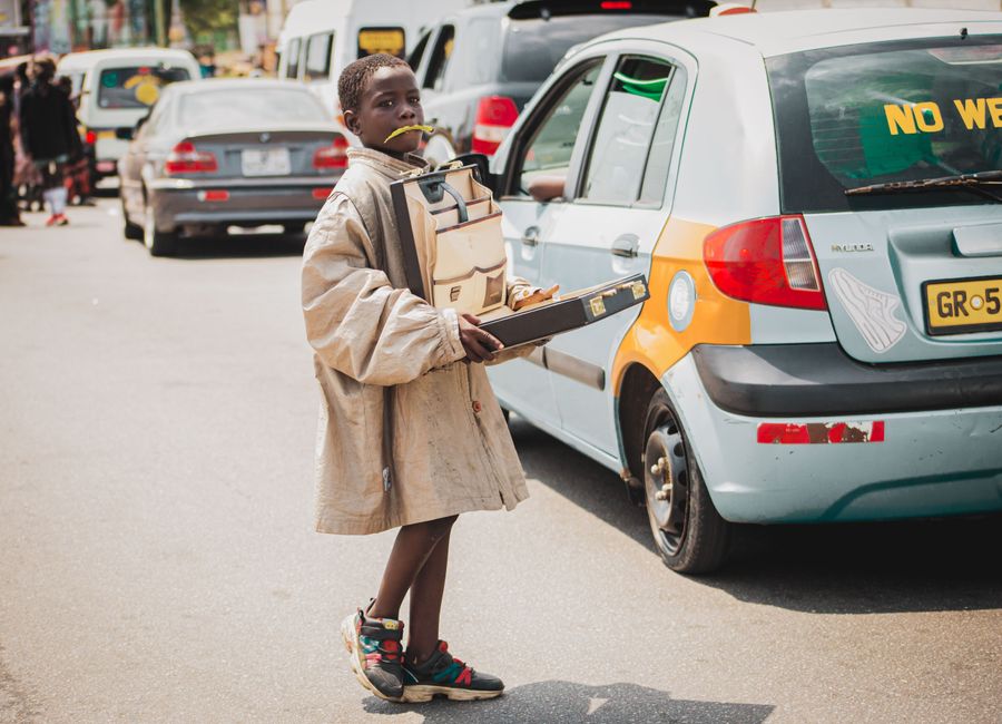 A young boy holds a box standing next to cars on a bustling street in Accra, Ghana.