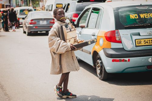 Free A young boy holds a box standing next to cars on a bustling street in Accra, Ghana. Stock Photo