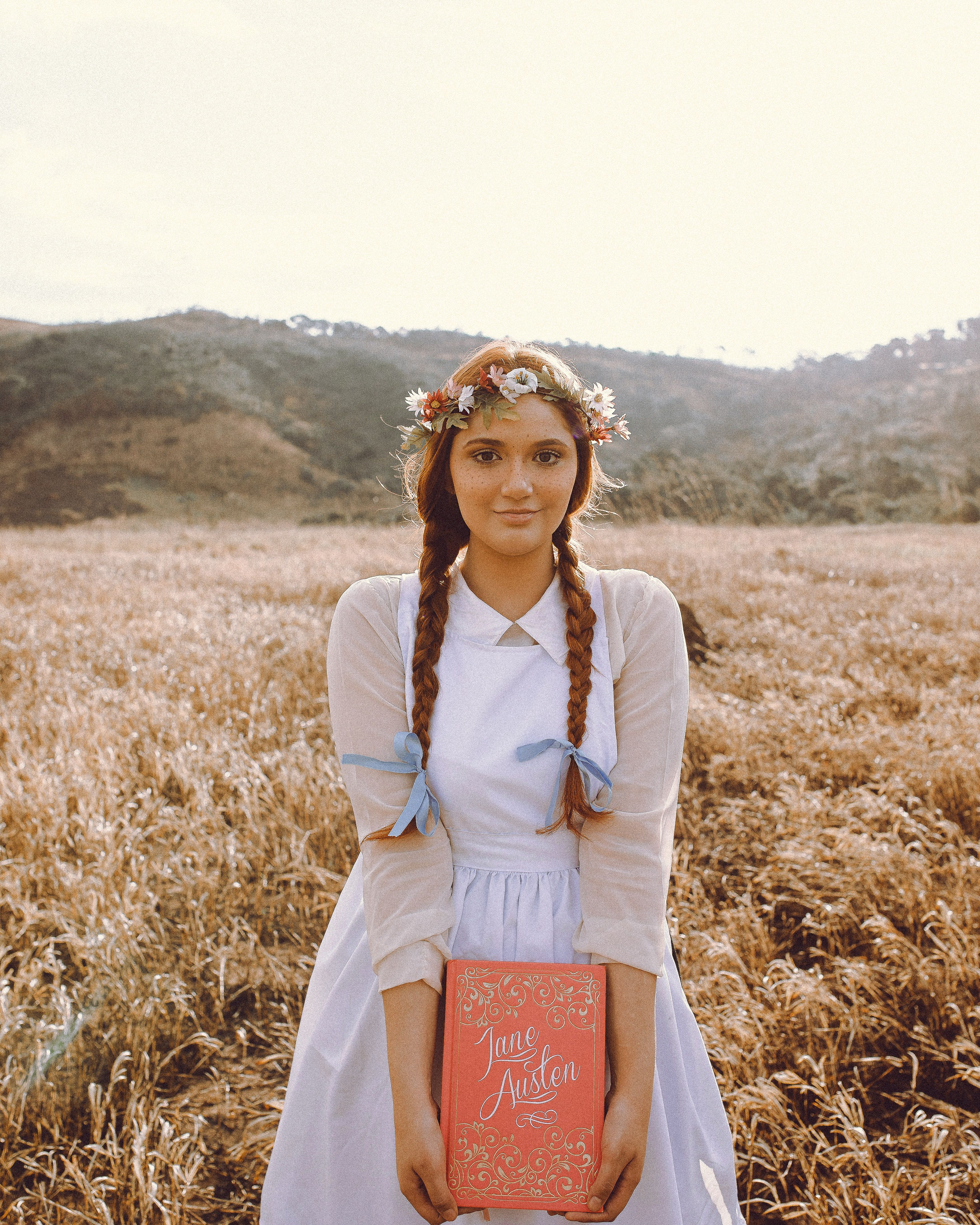 A young woman in a vintage dress holds a Jane Austen book in a sunlit field.