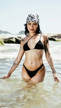 Confident woman in a black bikini with tattoos stands in the clear sea on a sunny day.