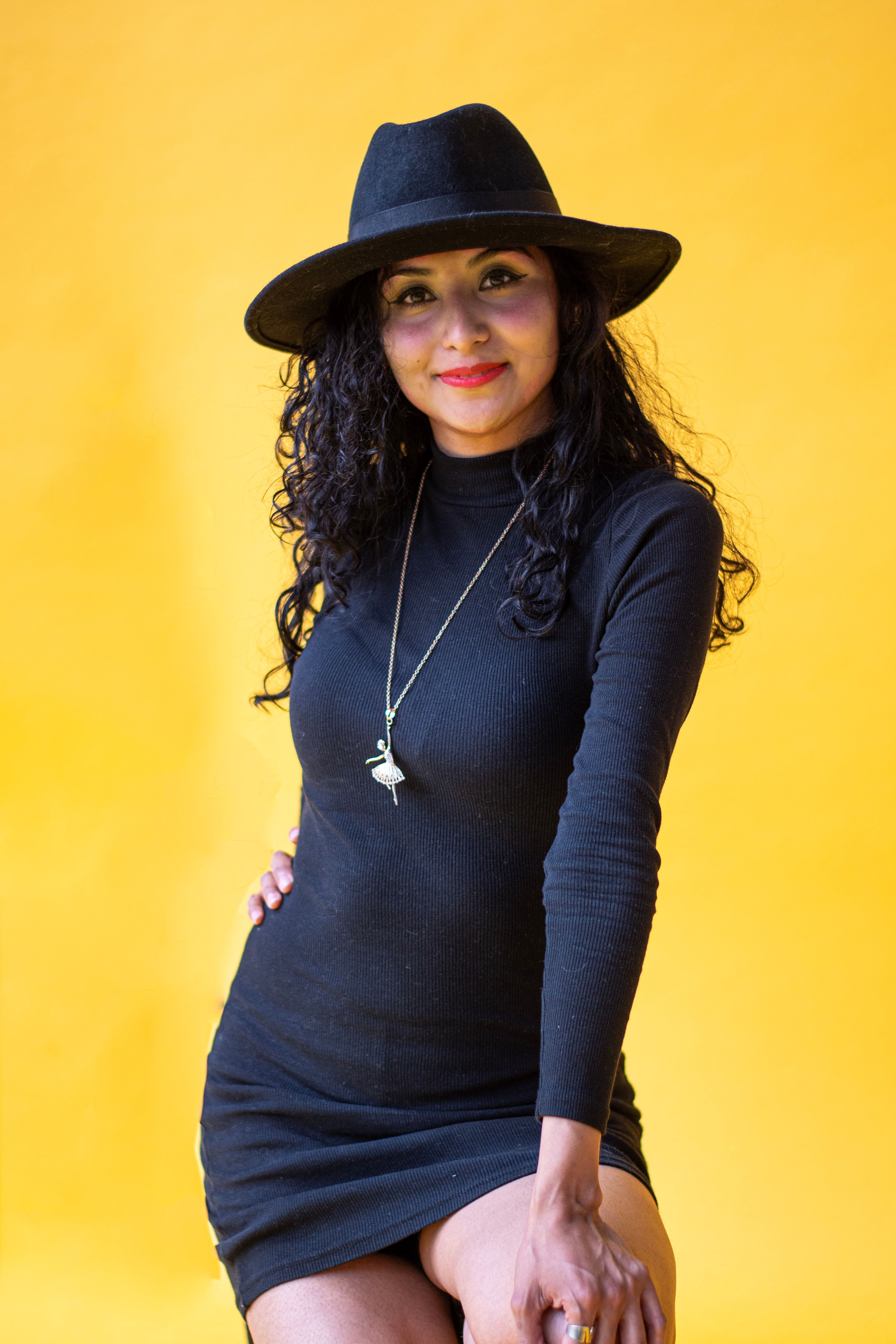 Fashionable woman posing confidently in black dress and hat against vibrant yellow background.