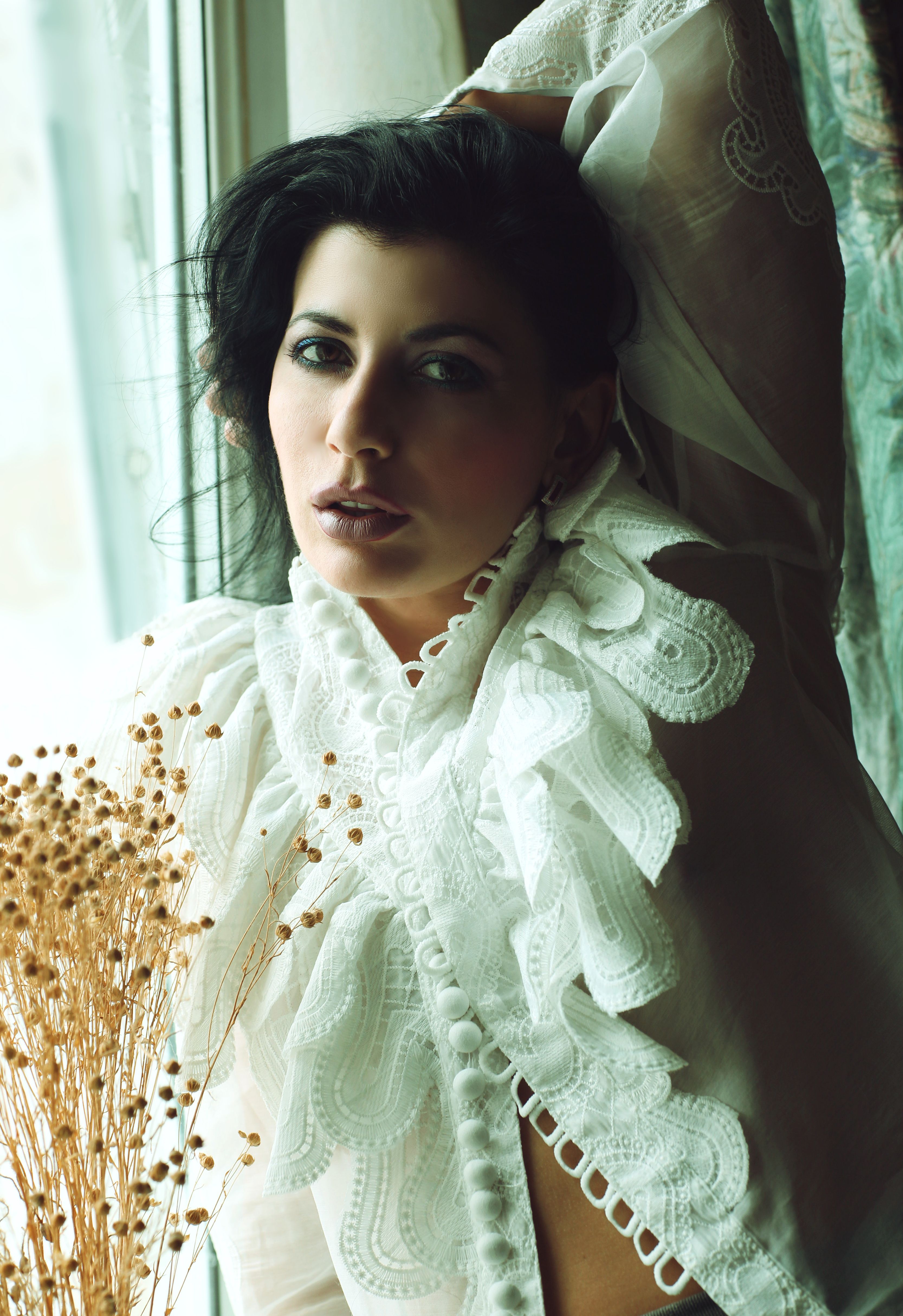 Fashion portrait of a woman with black hair in a white lace blouse by a window.