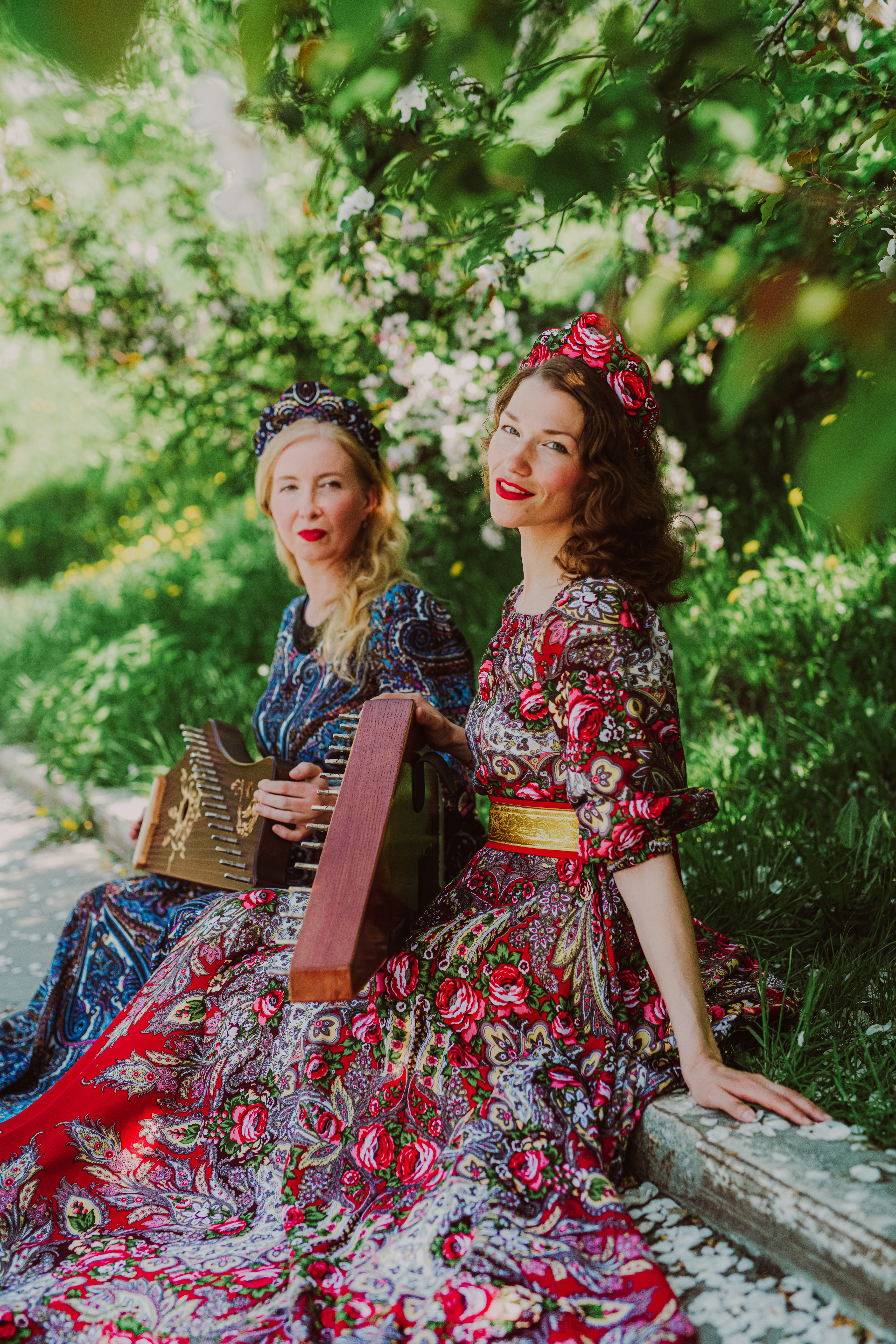 Two women in ornate floral dresses playing musical instruments in a sunny park setting.