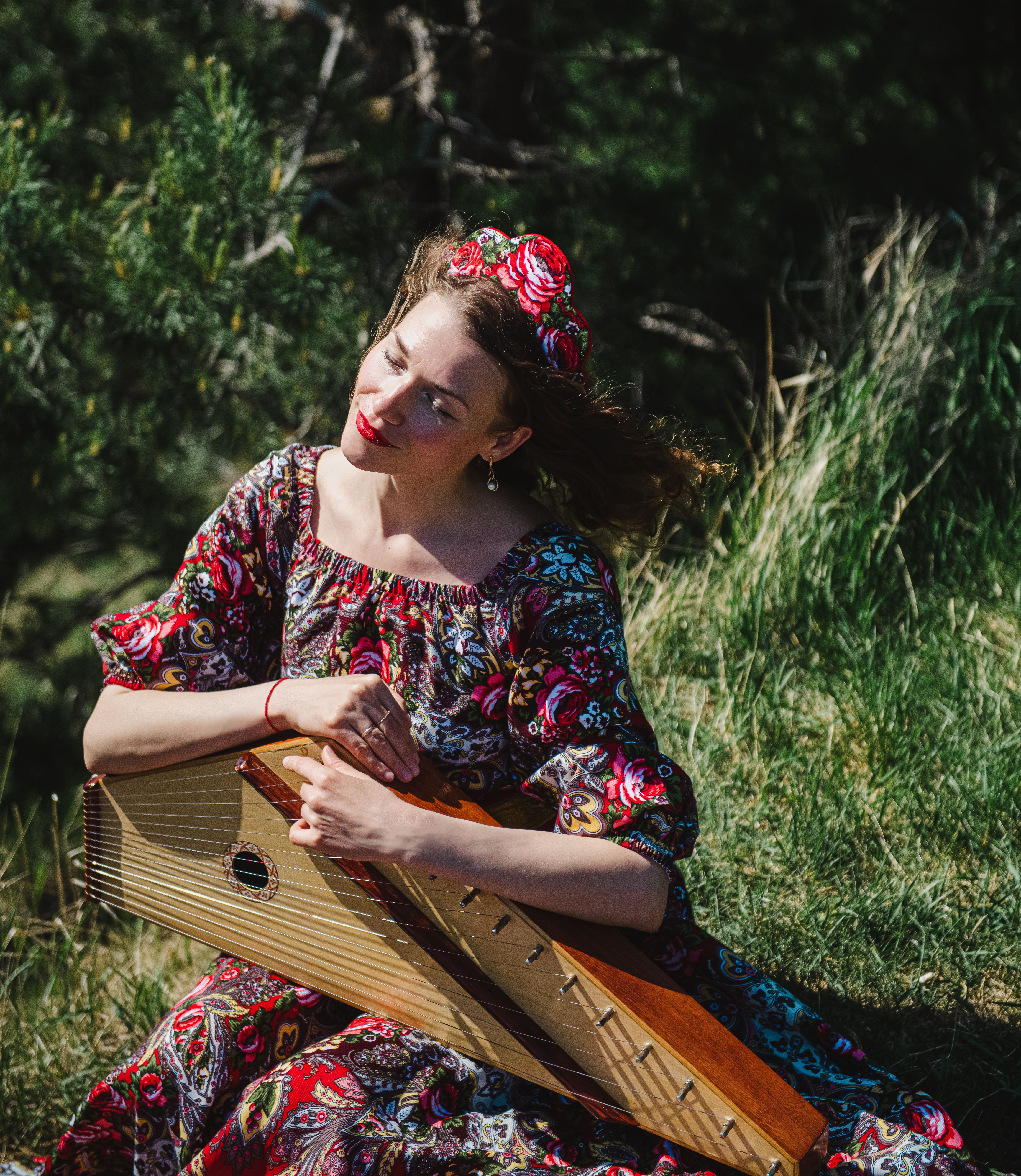 Woman in colorful dress plays folk instrument sitting outdoors with eyes closed, enjoying nature.