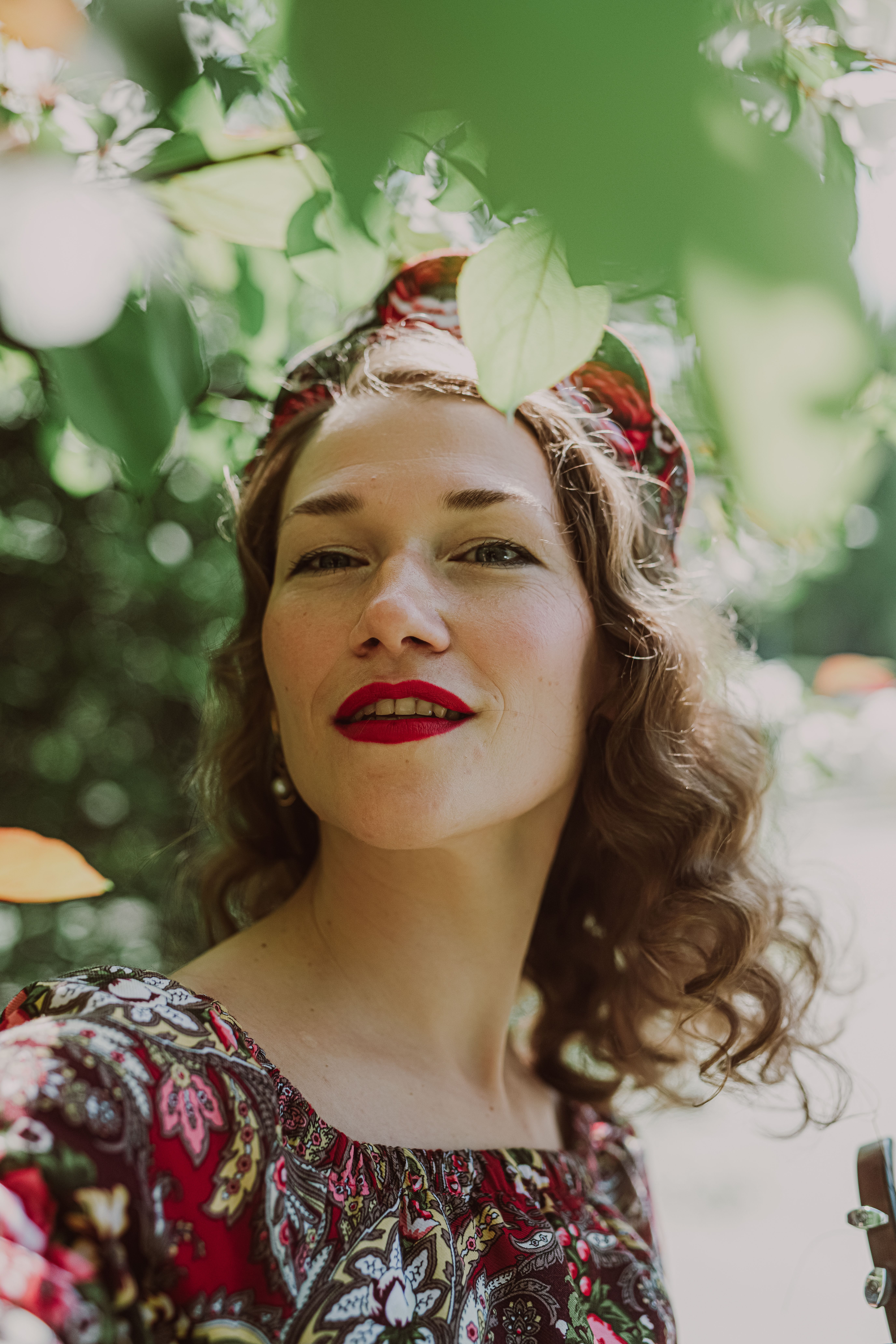 A stylish woman with long brown hair posing outdoors surrounded by lush greenery.