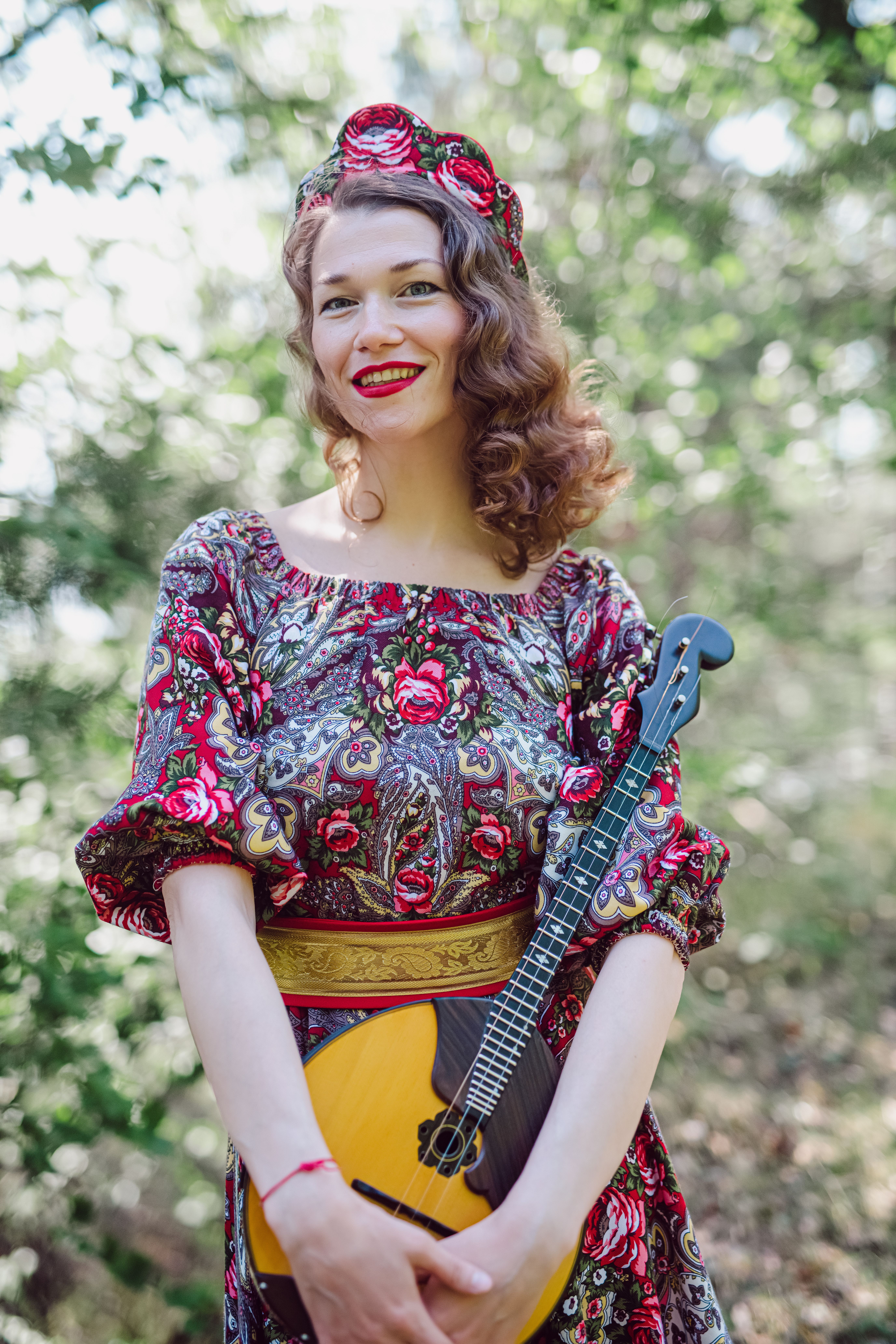 Caucasian woman in a floral dress holding a dombra and smiling outdoors.