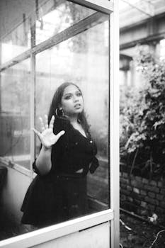 Moody black and white portrait of a woman standing indoors behind a rain-streaked window.