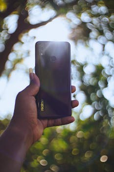 Close-up of hand holding smartphone with sunlight and blurred bokeh background, emphasizing technology in nature.