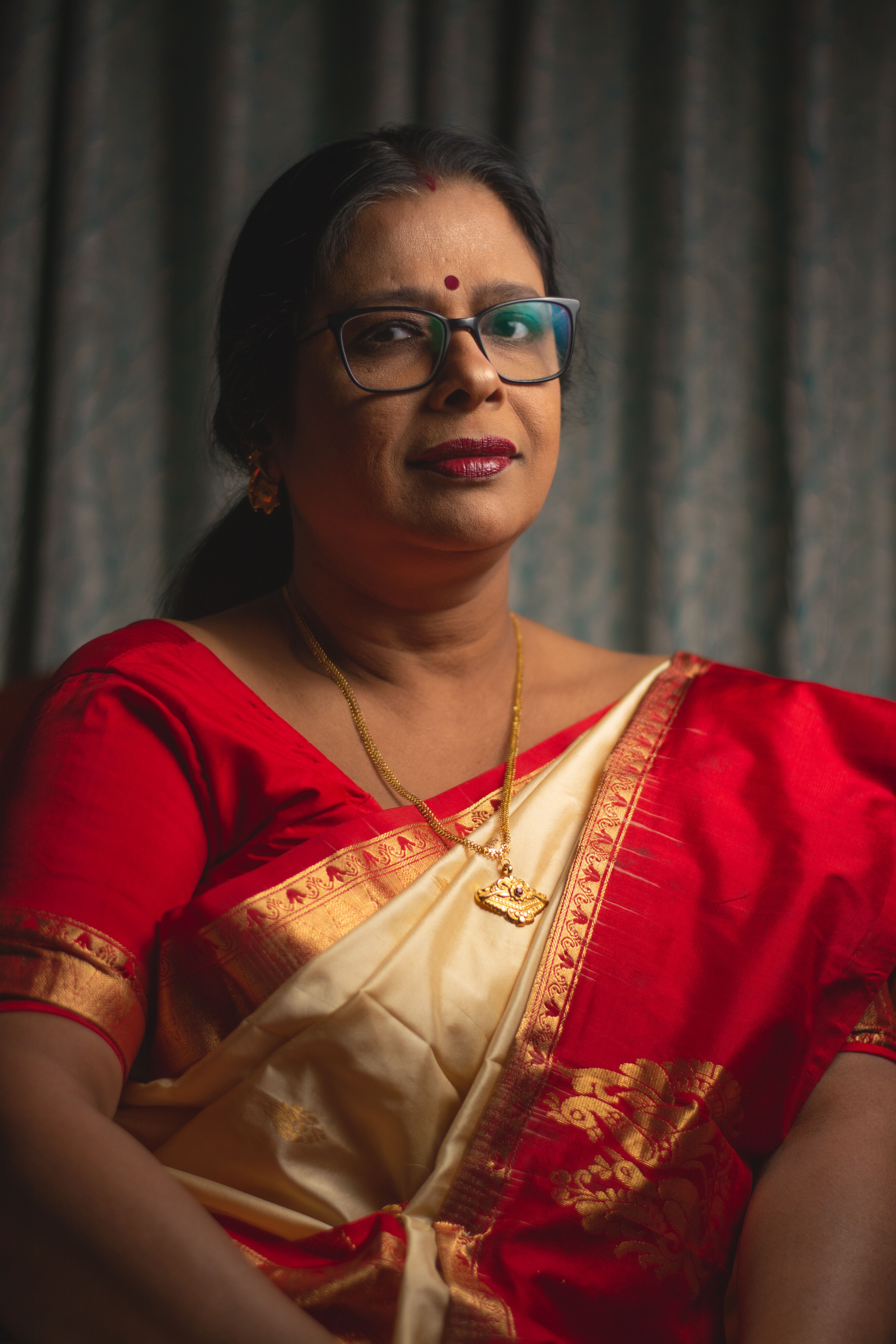 A woman in a vibrant red and gold saree, wearing eyeglasses and jewelry, poses inside.