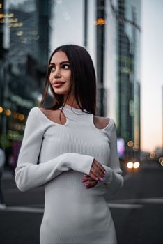 Stylish woman in white dress posing on a city street in twilight with skyscrapers in the background.