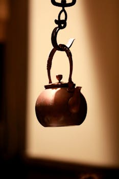 A beautifully crafted antique copper kettle hanging on a hook with warm sunlight casting dramatic shadows.