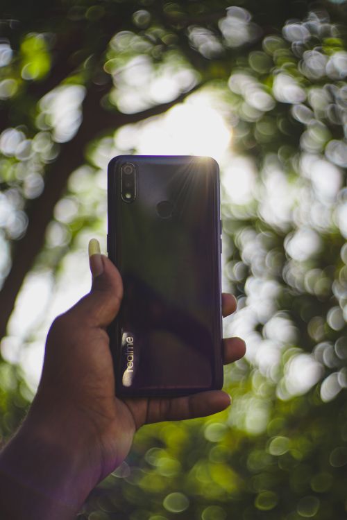 Free Close-up of a smartphone held by a hand with sunlight and bokeh background. Stock Photo