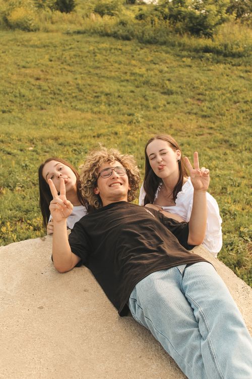 Free Three young friends lie on the grass, smiling and posing with peace signs in a sunny park setting. Stock Photo