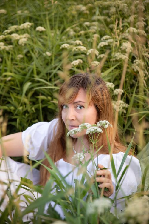 Free A woman in a white dress surrounded by lush green wildflowers, capturing summer's essence. Stock Photo