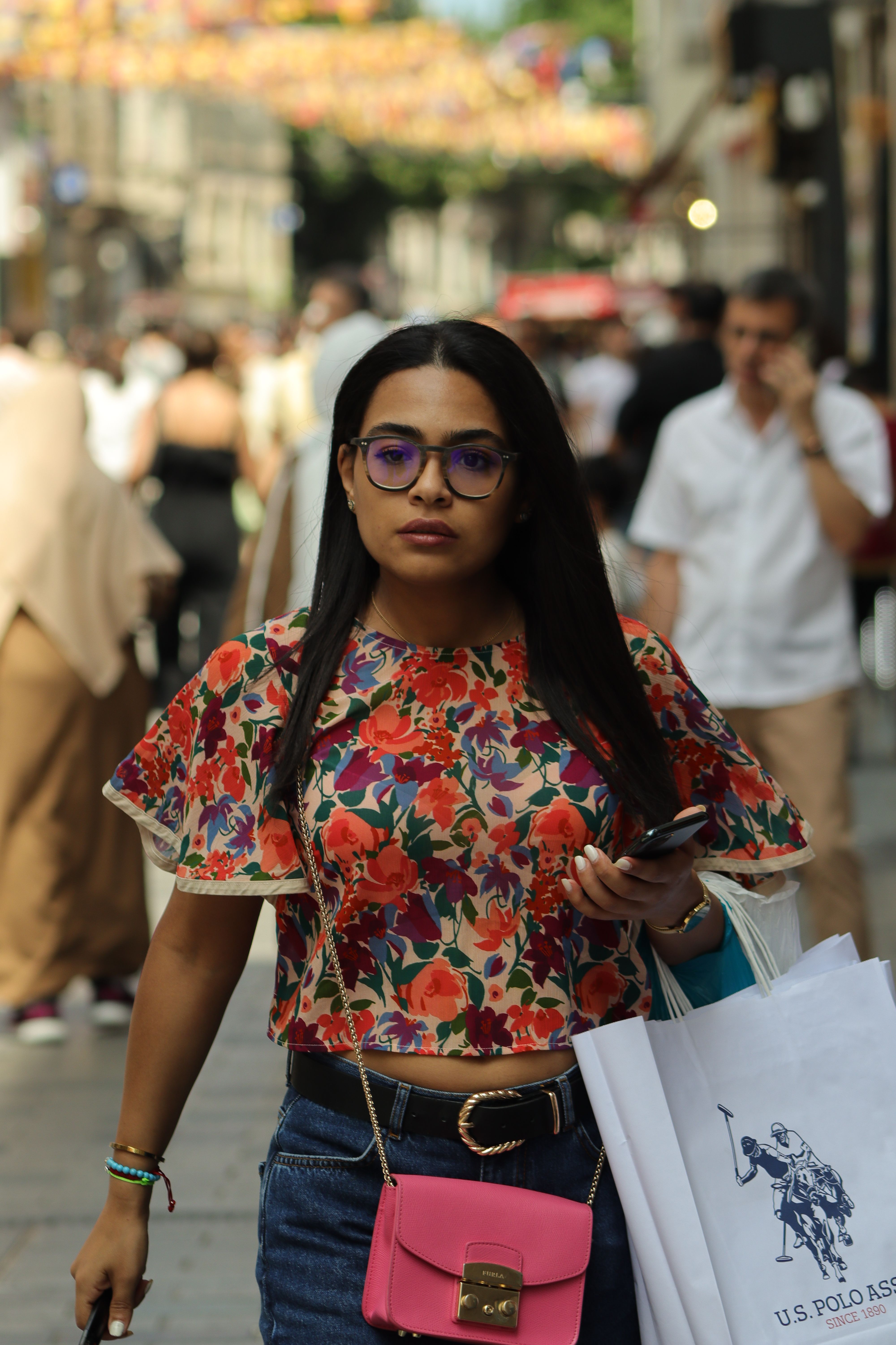 Young woman in a floral blouse walking with shopping bags on a bustling urban street. - Care and Maintenance Tips for Handbags to Prolong Their Life