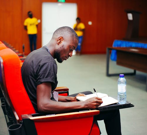 Free Black man sitting in a conference room, writing notes with a water bottle visible. Stock Photo
