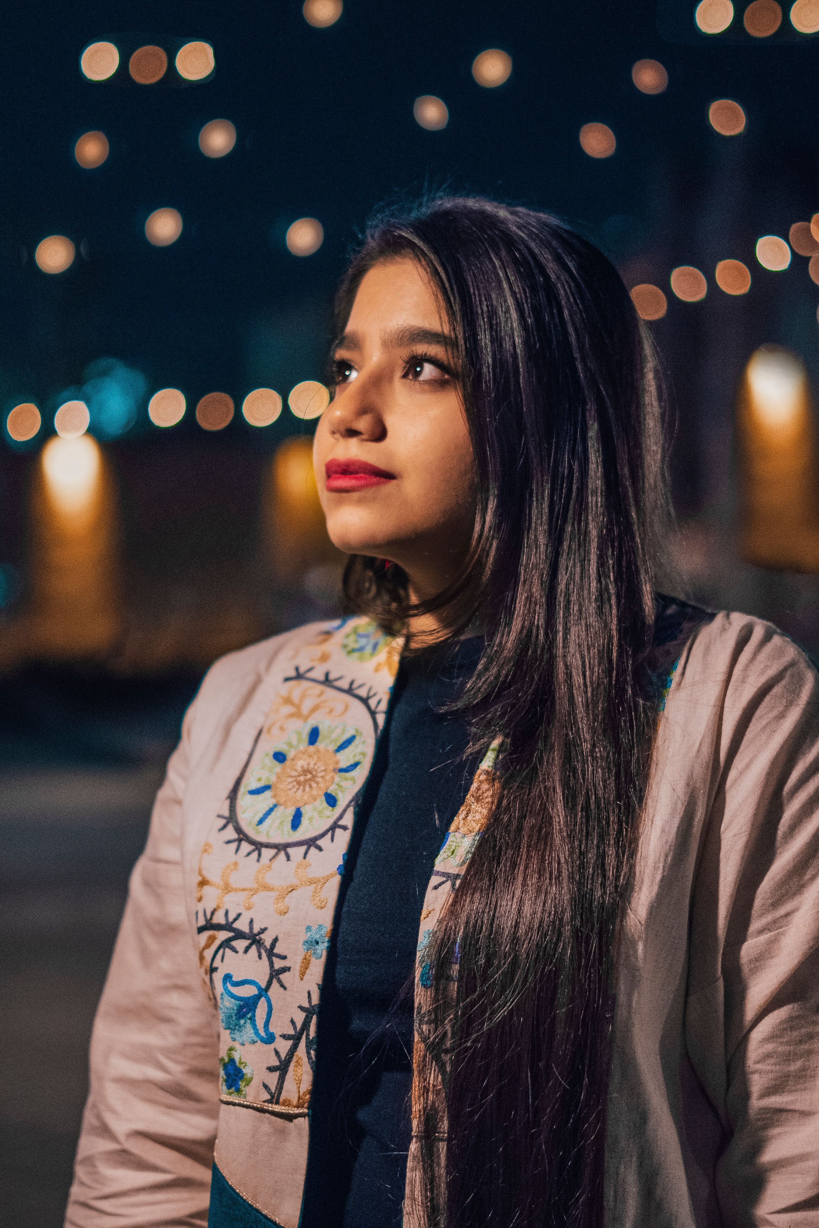 A woman with long hair wearing an embroidered jacket looks upwards, captured at night with bokeh lights.