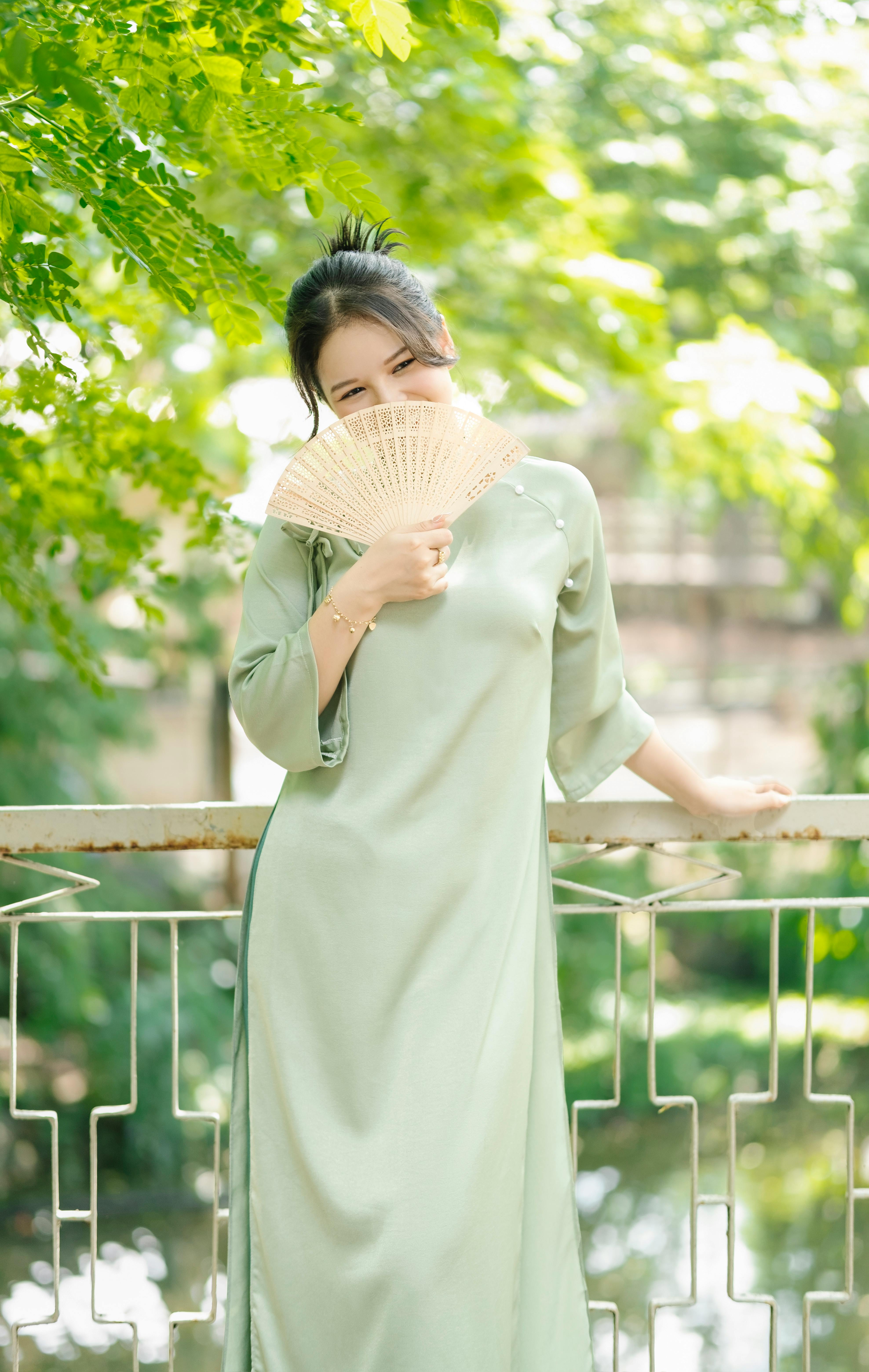 Sophisticated woman in a green dress holds a folding fan on a sunny day outdoors.