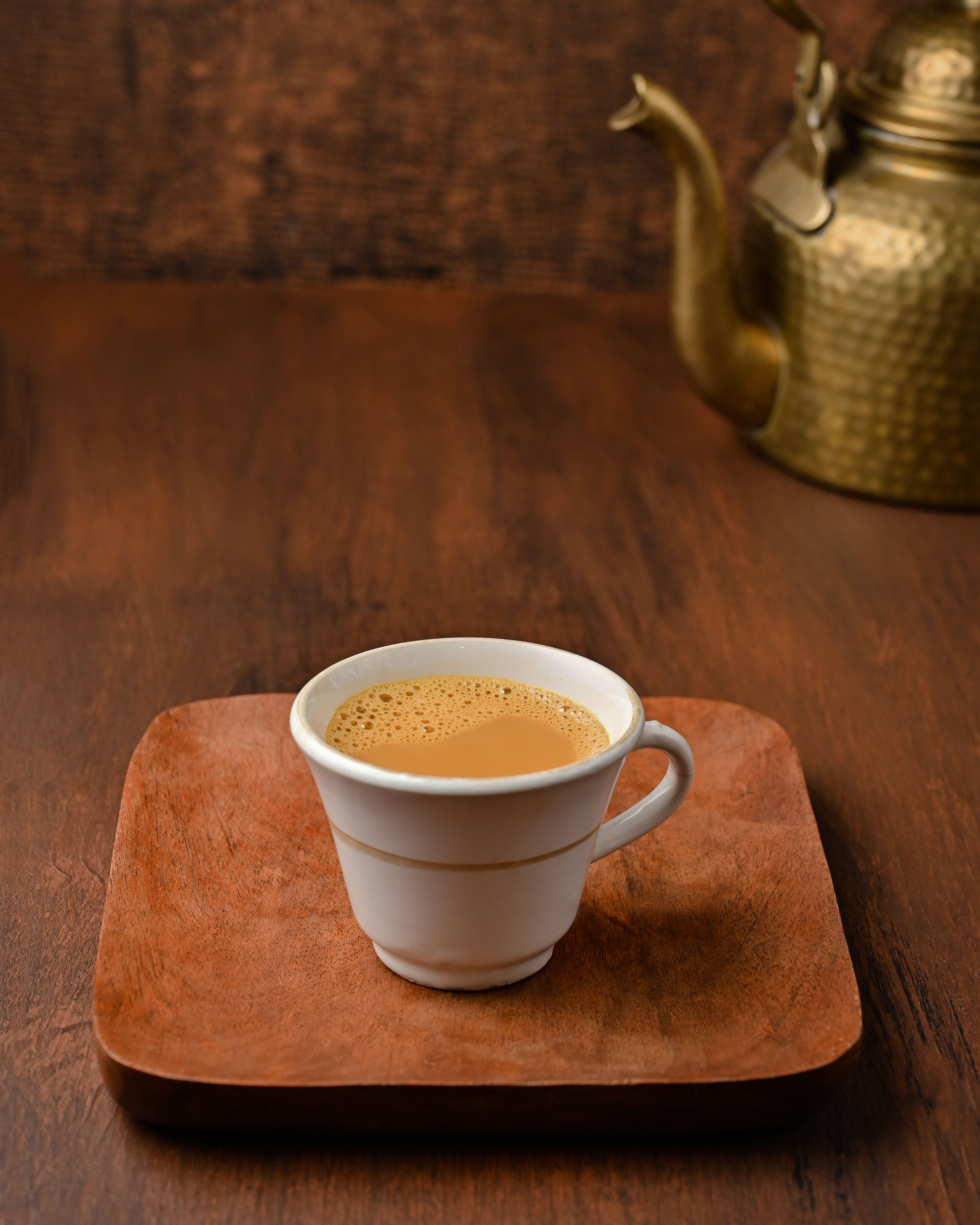 A cup of traditional Indian tea served on a wooden tray beside a brass kettle.