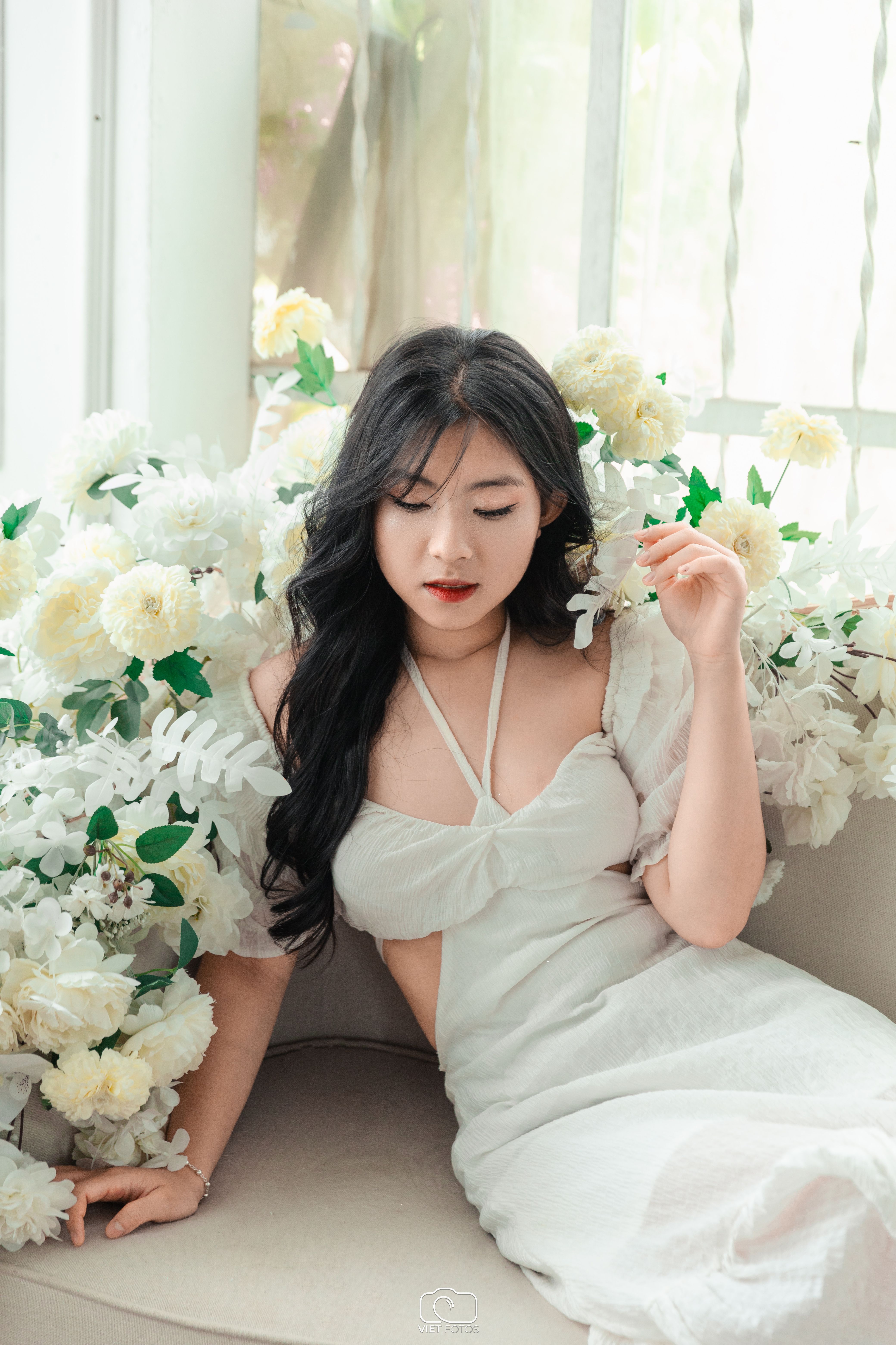 A young Asian woman elegantly posing in a white dress amidst a beautiful flower arrangement.