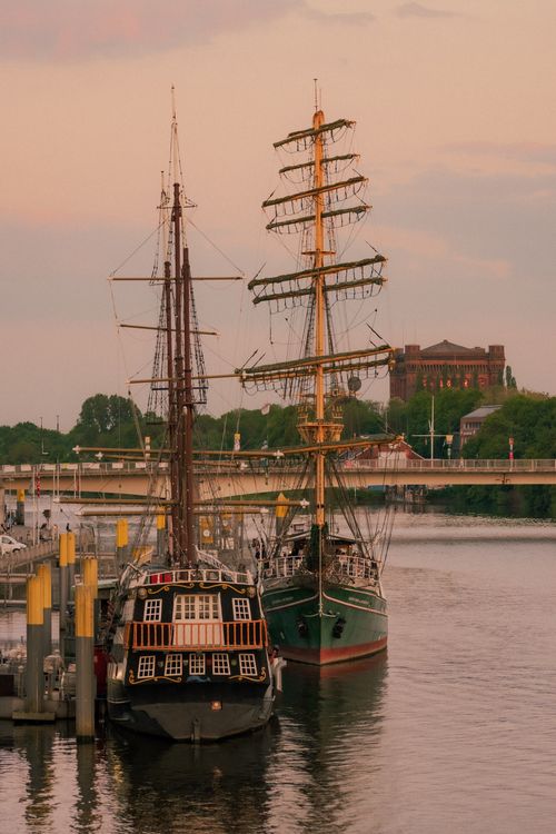 Free Historic sailing ships moored by a river at sunset with a bridge and vintage architecture. Stock Photo