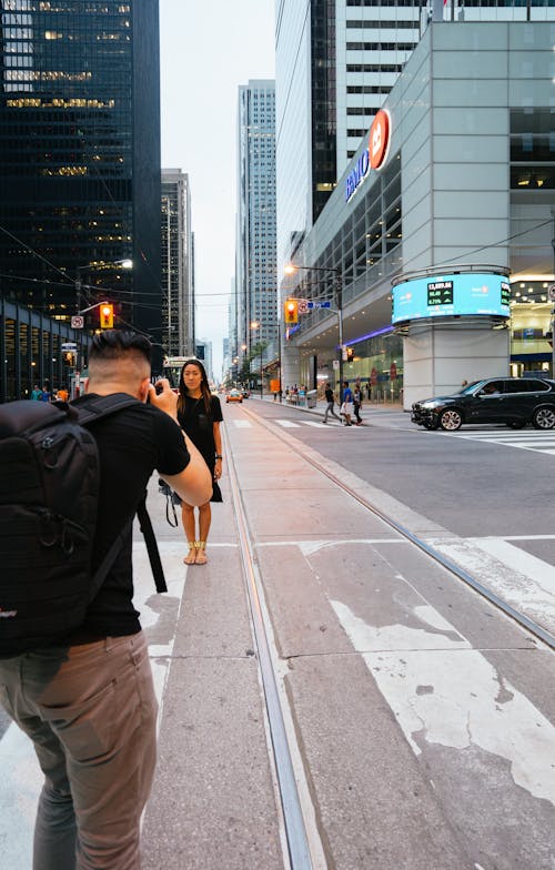 Man Taking Photo of Woman Standing Beside Highway Near Building