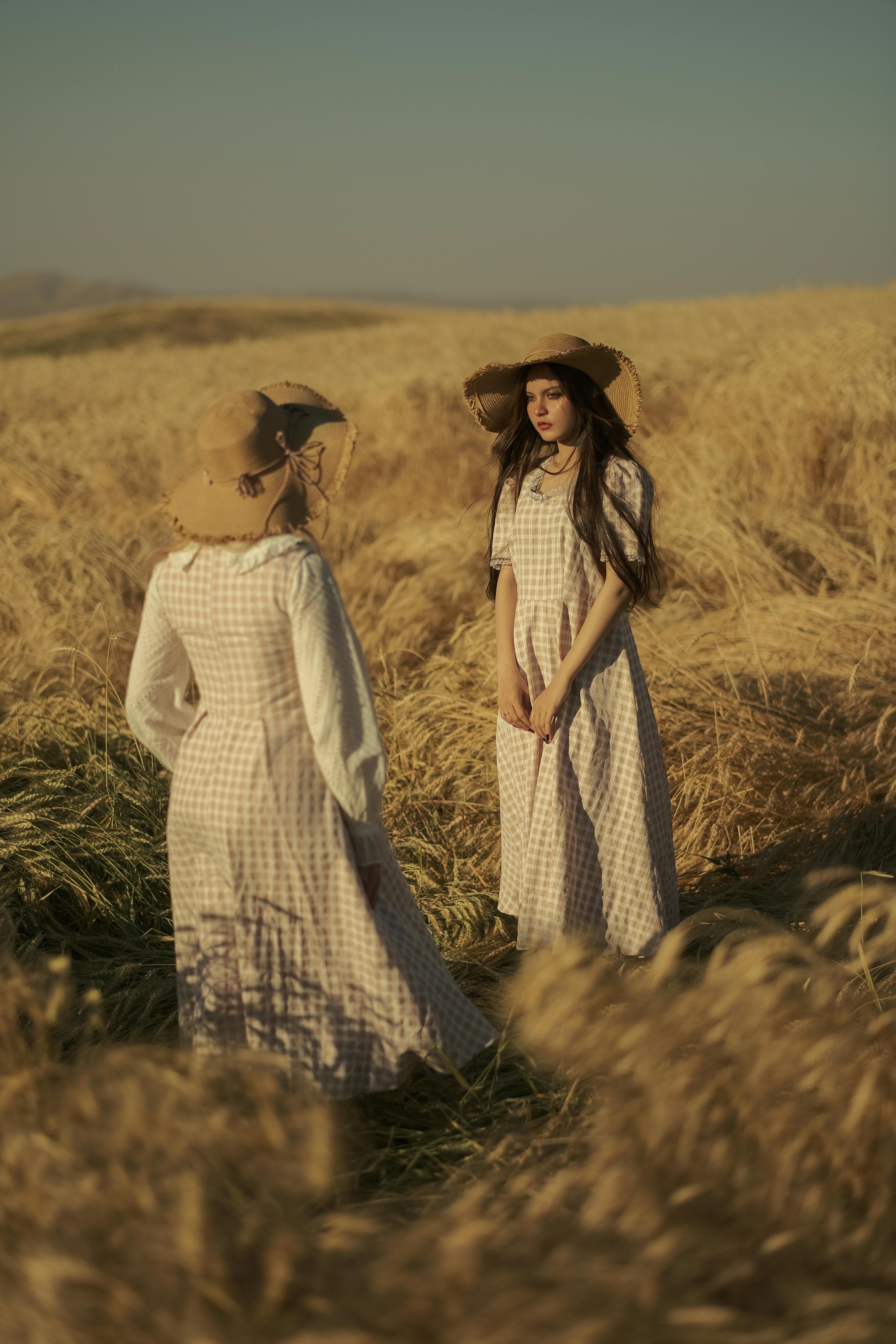 Two women in long dresses and hats stand in a sunlit wheat field, capturing a summer fashion vibe.