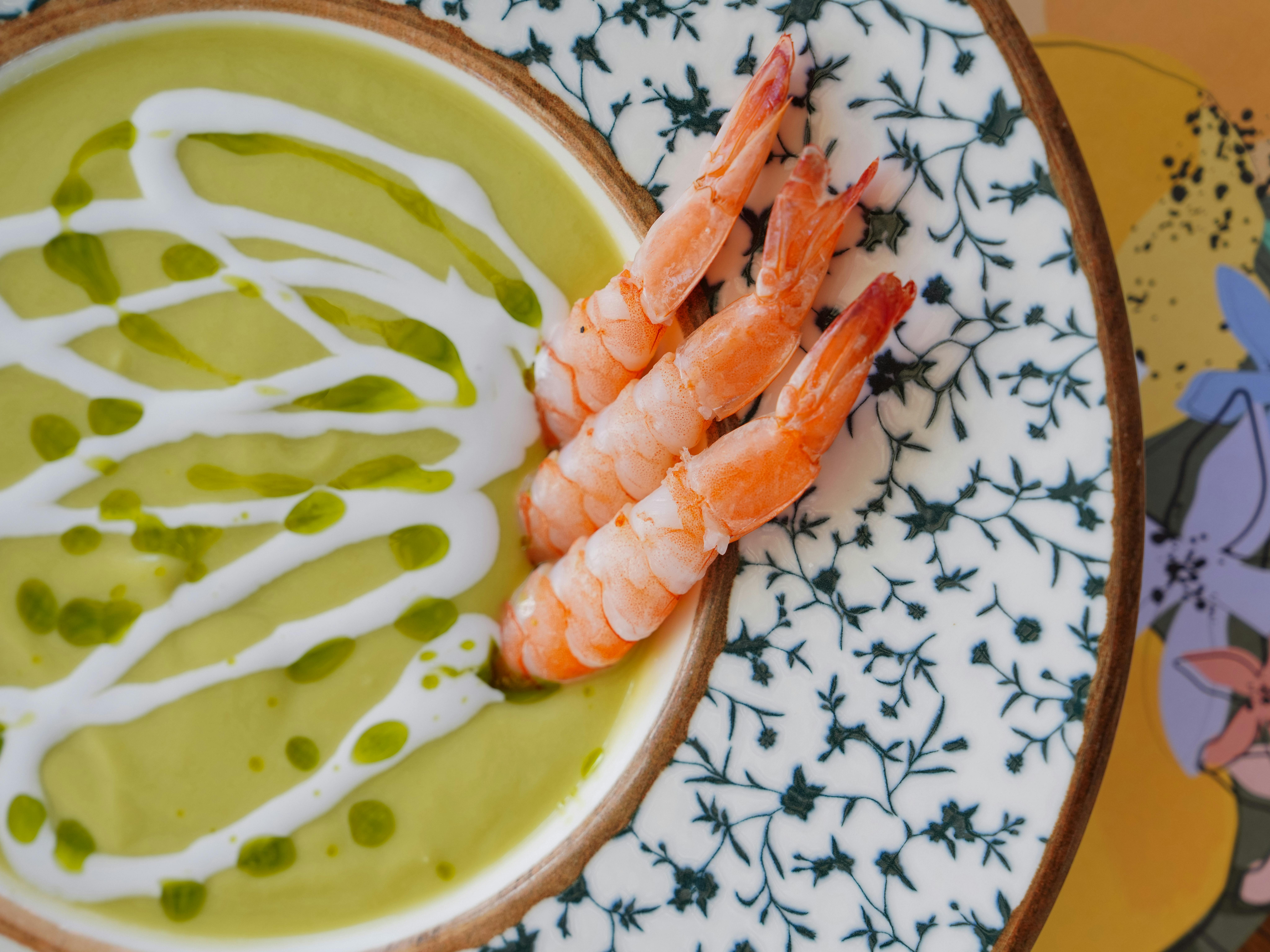 A close-up shot of shrimp served on avocado soup in a decorative bowl, garnished with cream and herbs.