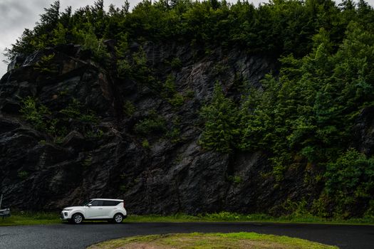 A white compact car on a rain-soaked road beside a lush, green forested rock face.