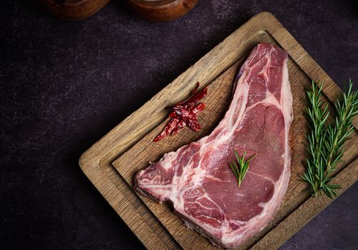 Top view of a raw steak on a cutting board with rosemary and red chili peppers.